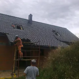 Montaż czarnej dachówki na nowym domu z cegły. Na rusztowaniu dekarz w trakcie pracy, drugi obserwuje z dołu. Widoczne okna dachowe i komin. Pochmurne niebo w tle.