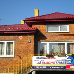 Ceglany dom z czerwonym dachem z blachy trapezowej, żółte rynny, reklama firmy Blachotrapez na balkonie, błękitne niebo.