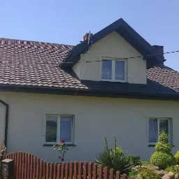 Dom jednorodzinny z brązowym dachem z dachówki ceramicznej, czarnymi rynnami i białą elewacją, otoczony zielenią i niskim płotem z drewna.