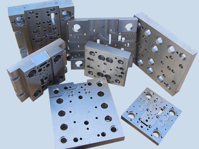 Zestaw metalowych form wtryskowych o różnych rozmiarach i kształtach, z widocznymi otworami i frezowaniami, ułożonych na jasnym tle. Widoczne ślady obróbki.