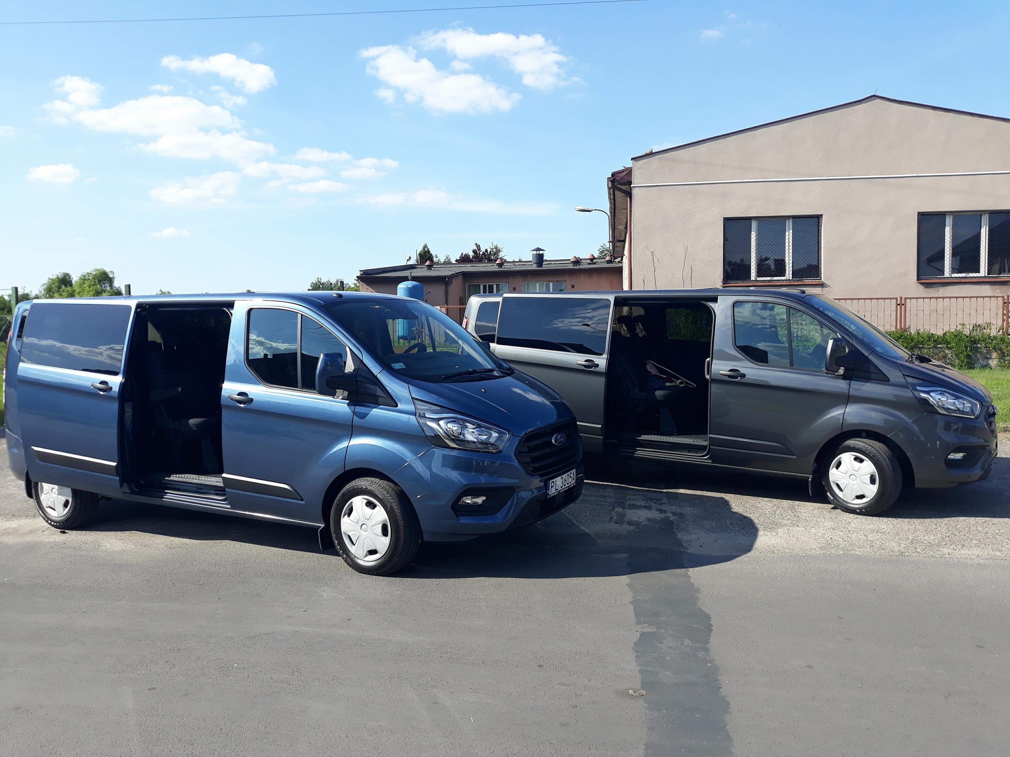 Dwa busy Ford Transit Custom, jeden niebieski z otwartymi drzwiami bocznymi, drugi szary z częściowo otwartymi drzwiami, stojące na asfalcie przed budynkiem w słoneczny dzień.
