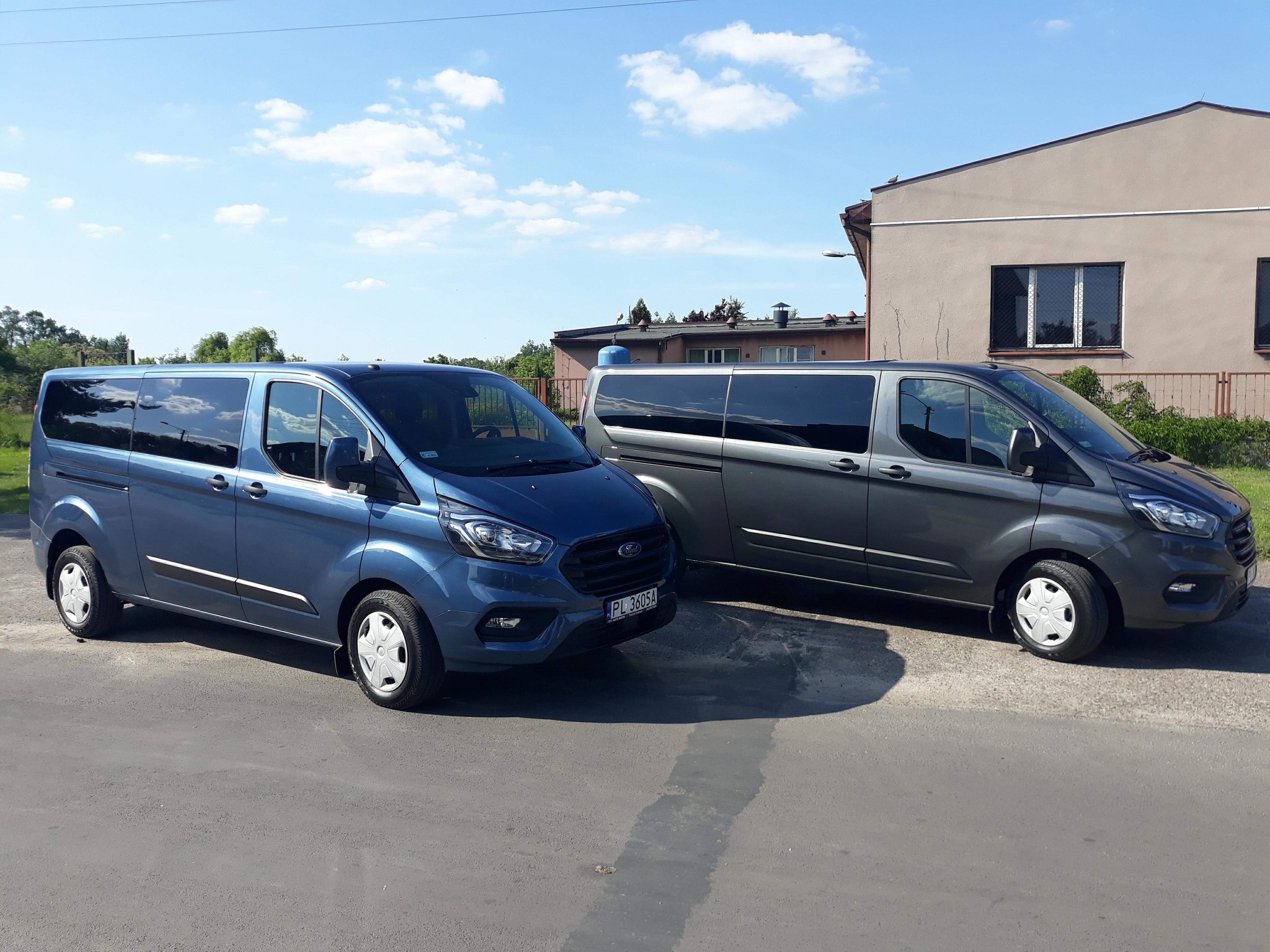 Dwa busy Ford Transit Custom w kolorze niebieskim i szarym zaparkowane obok budynku z kratami w oknach, widoczne tablice rejestracyjne PL.
