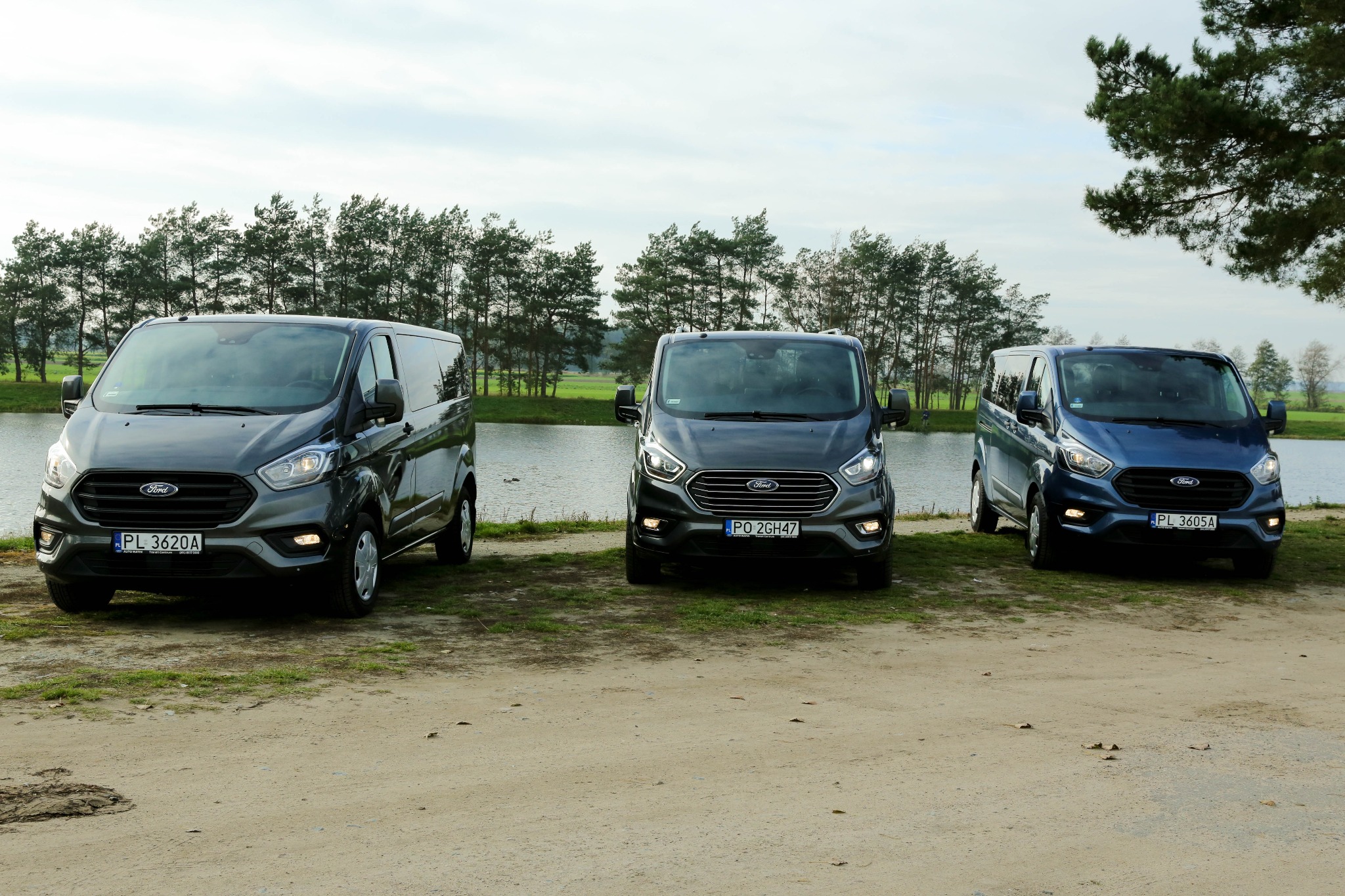 Trzy busy Ford Transit Custom w różnych kolorach (szary, ciemnoszary i niebieski) stojące obok siebie na trawiastym brzegu jeziora, z widocznymi tablicami rejestracyjnymi.
