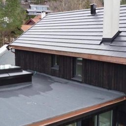 Jakub Lipka - Nowoczesny dach z ciemnej dachówki ceramicznej i jasnym kominem, wykończony miedzianymi obróbkami blacharskimi. Widok z góry na dom z płaskim dachem i świetlikiem.