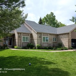 Jakub Lipka - Dom z szarego, nowego dachu z ceglaną elewacją, zadbanym trawnikiem i elementami zieleni. Widoczne okna z białymi okiennicami i garaż z samochodem w środku.