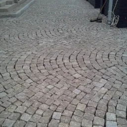 Ułożona z kostki granitowej ścieżka z wzorem wachlarzowym, widok z góry. Na skraju widoczny fragment budynku i narzędzia brukarskie.