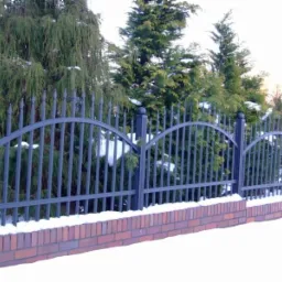Ciemnoszare przęsło ogrodzeniowe z metalowymi sztachetami i łukowym wykończeniem, osadzone na murku z czerwonej cegły, w zimowej scenerii z iglakami w tle i śniegiem na gruncie.