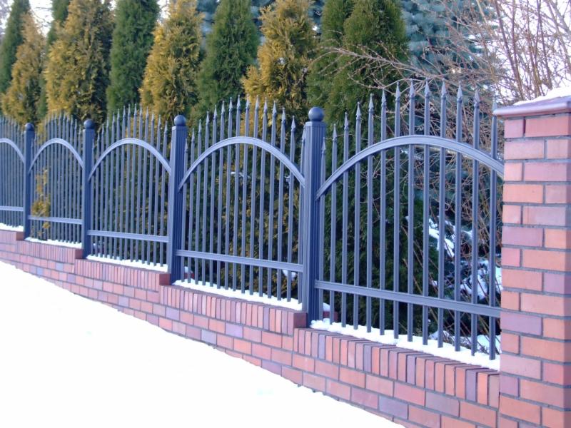 Nowoczesne przęsło z metalowych sztachet, łukowato zakończone, osadzone na niskim murku z czerwonej cegły, w tle zimozielone drzewa i śnieg.