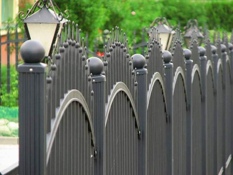 Czarne, metalowe przęsła ogrodzeniowe z dekoracyjnymi łukami i kulami na słupkach, oświetlone latarniami, na tle zieleni.