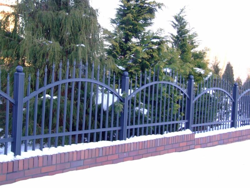 Ciemnoszare przęsło ogrodzeniowe z metalowymi sztachetami i łukowym wykończeniem, osadzone na murku z czerwonej cegły, w zimowej scenerii z iglakami w tle i śniegiem na gruncie.