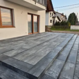 Nowoczesny taras z szarych płyt kamiennych, z widocznymi schodami prowadzącymi do domu z balkonem, w tle zabudowa jednorodzinna.