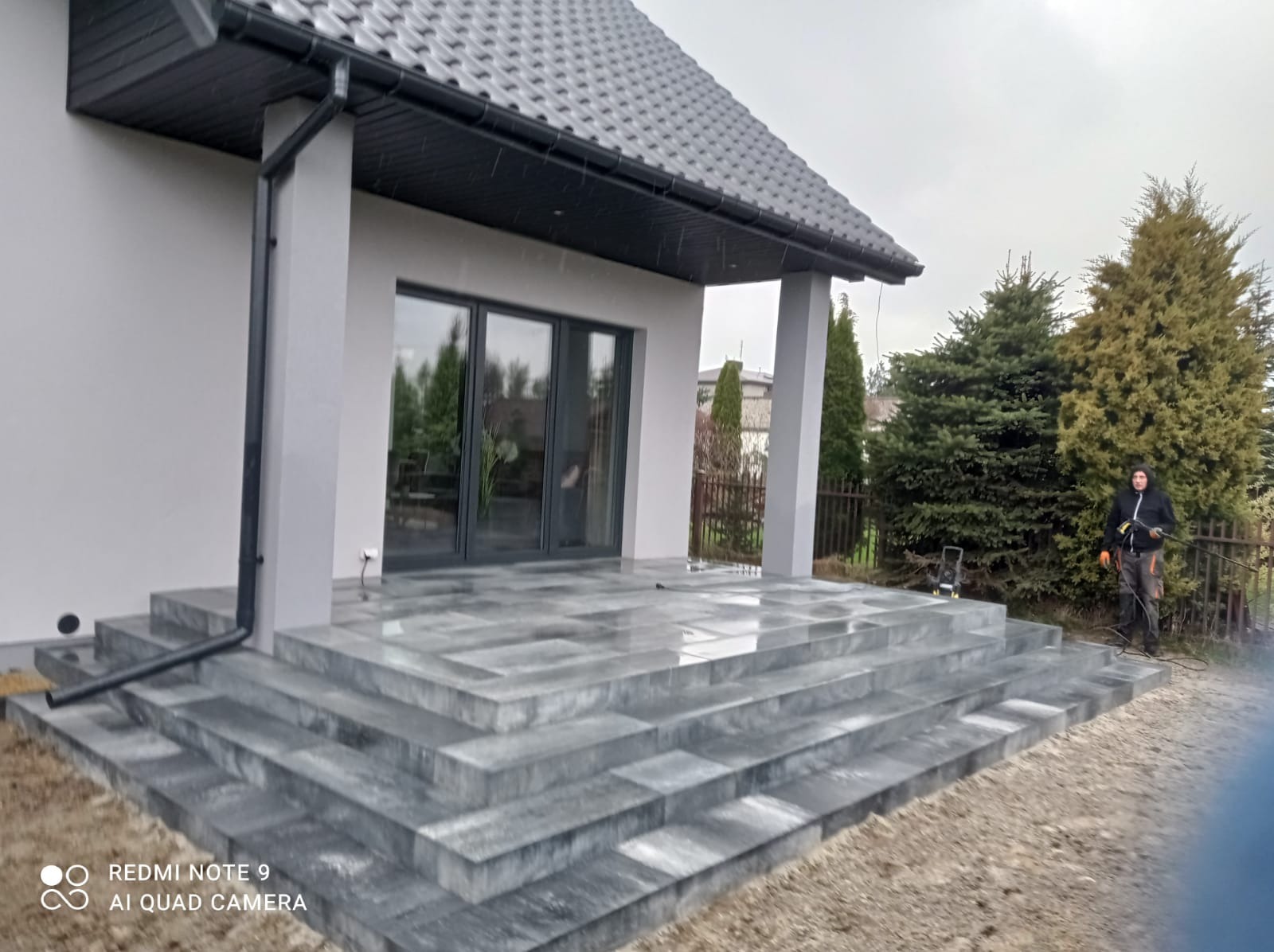Nowoczesny taras z szarej kostki brukowej prowadzący do domu z dużymi, przesuwnymi drzwiami, widoczny pracownik z myjką ciśnieniową w tle.
