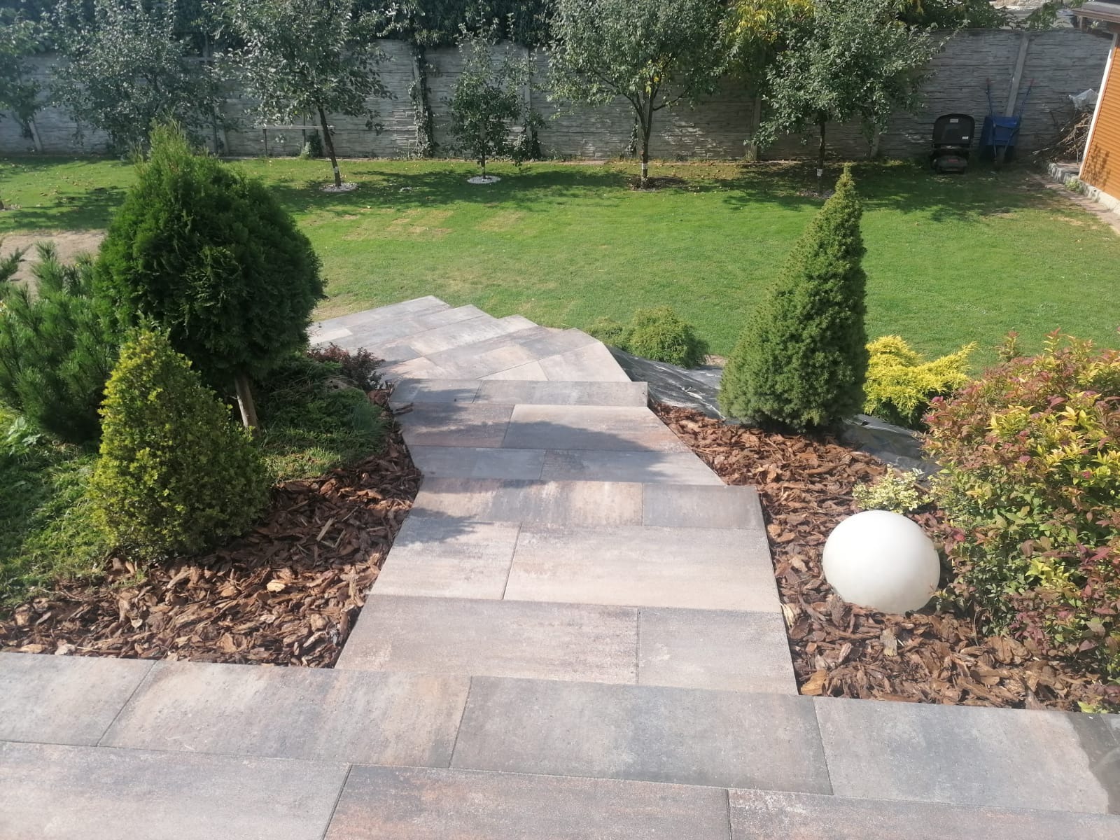Schody z kostki brukowej prowadzące w dół ogrodu, otoczone korą, iglakami i krzewami ozdobnymi, z dekoracyjną kulą oświetleniową.
