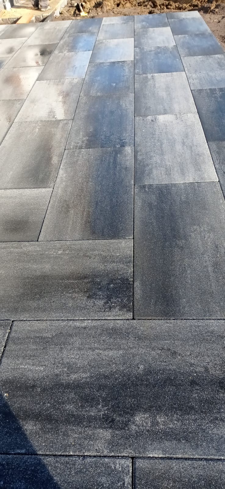 Nowo ułożona, szara kostka brukowa o prostokątnym kształcie, częściowo mokra, odbijająca światło, widoczna krawędź terenu z ziemią.