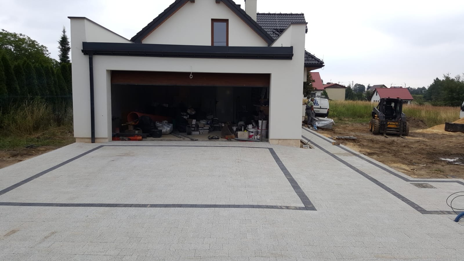 Jasnoszara kostka brukowa z ciemnoszarą obwódką przed garażem nowoczesnego domu; w tle miniładowarka i częściowo otwarty garaż z narzędziami i materiałami budowlanymi.