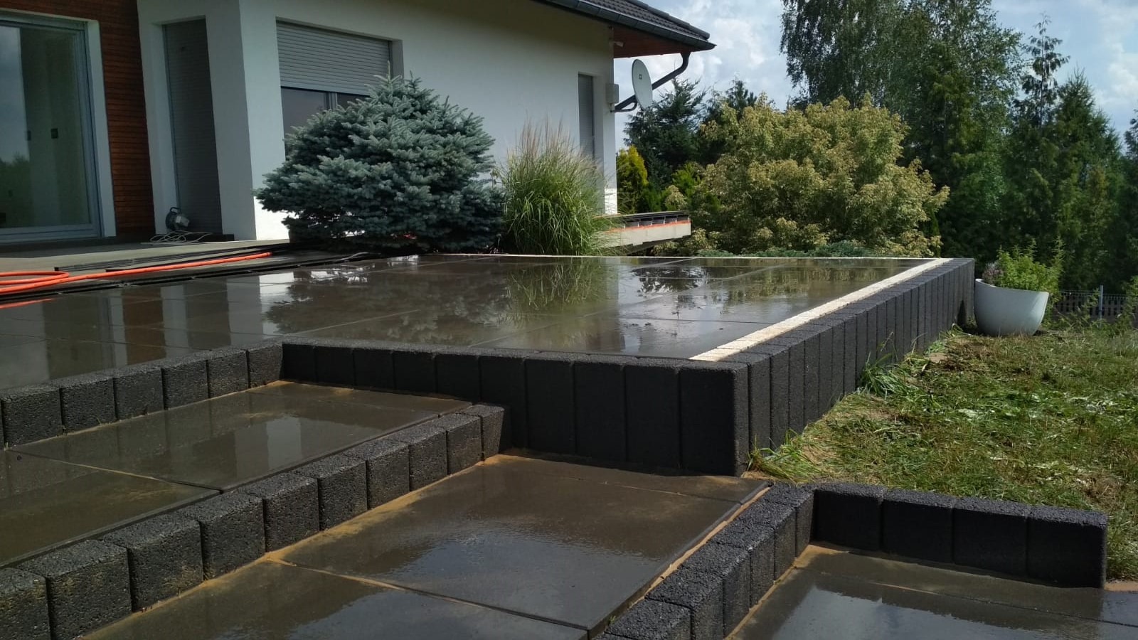 Nowoczesny taras z ciemnej kostki brukowej po deszczu, z niskimi schodami i obrzeżem, widok na ogród i biały dom z szarą stolarką okienną.