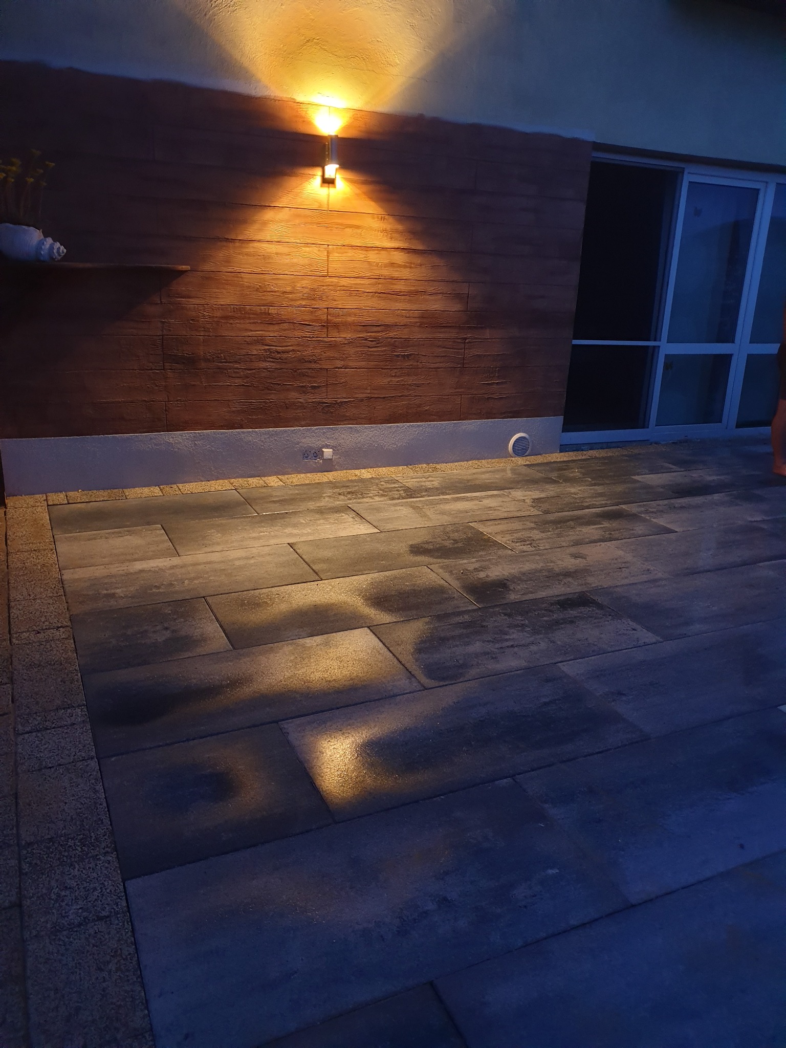 Nowoczesny taras z szarych, prostokątnych płyt, oświetlony lampą ścienną i subtelnym światłem pod ścianą, z elementami dekoracyjnymi na półce.