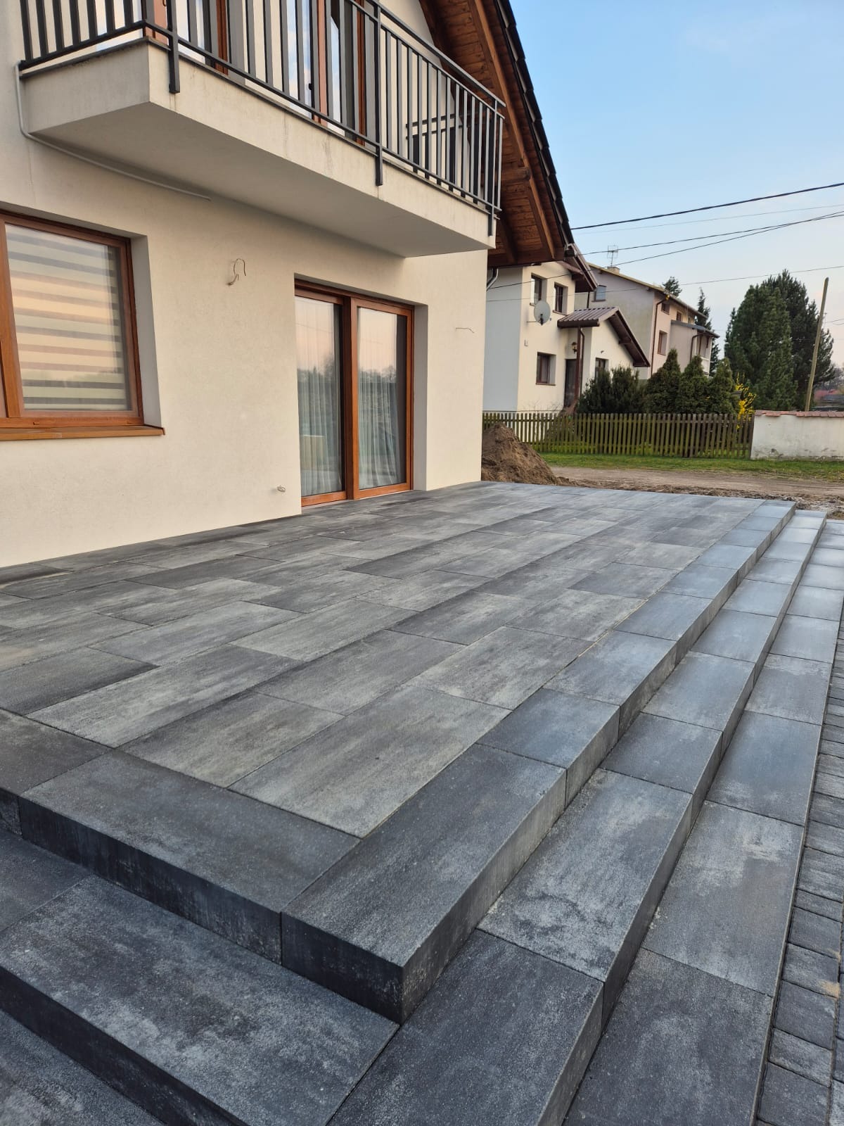 Nowoczesny taras z szarych płyt kamiennych, z widocznymi schodami prowadzącymi do domu z balkonem, w tle zabudowa jednorodzinna.