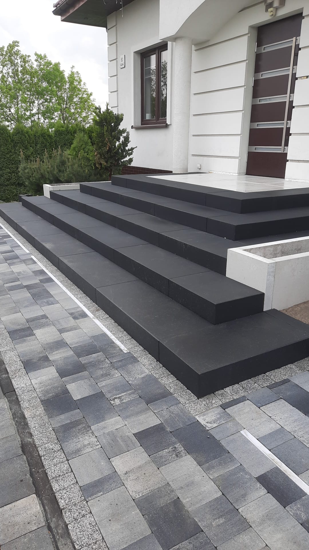 Nowoczesne schody wejściowe z ciemnoszarych płyt, harmonizujące z szaro-grafitową kostką brukową i jasną elewacją domu z nowoczesnymi drzwiami.