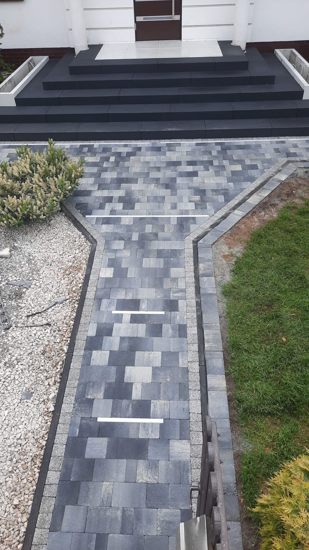 Ułożona ścieżka z kostki brukowej w odcieniach szarości, z białymi paskami odblaskowymi, prowadząca do schodów wejściowych, obok trawnik i rabata z kamykami.