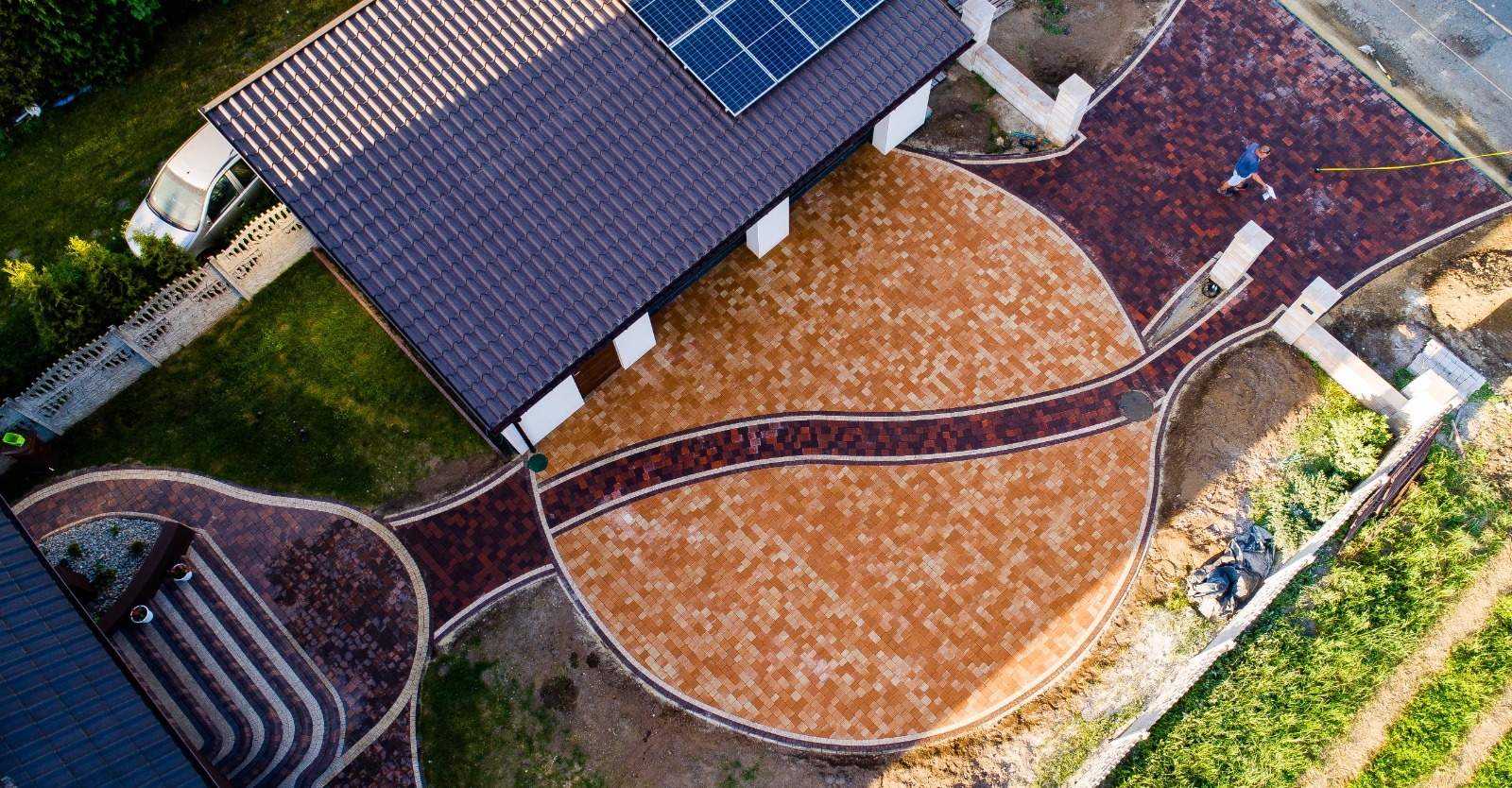 Widok z góry na posesję z nową, wzorzystą nawierzchnią z kostki brukowej w odcieniach beżu i brązu, z elementami dekoracyjnymi i pracownikiem z żółtą miarką, obok domu z panelami słonecznymi.