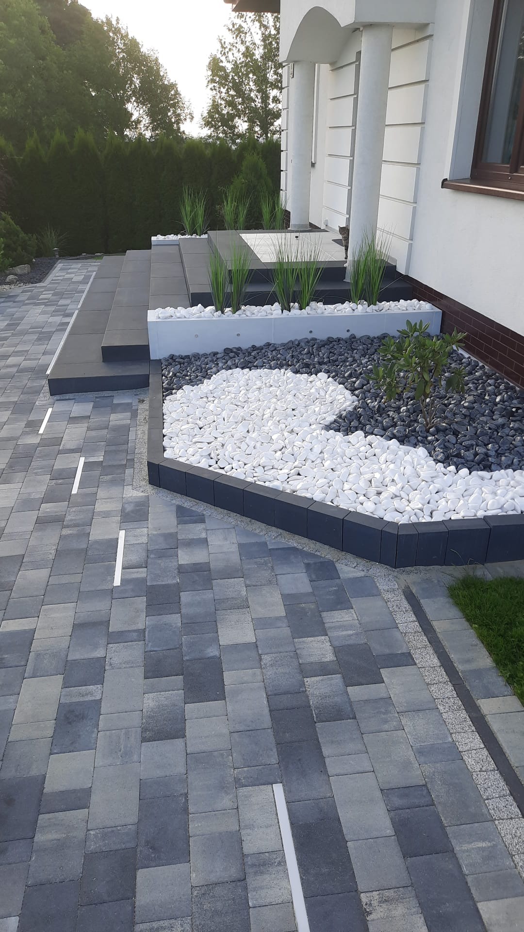 Ułożona kostka brukowa w odcieniach szarości prowadząca do schodów wejściowych z dekoracyjnym klombem z białych i szarych kamieni, oraz traw ozdobnych i krzewu.