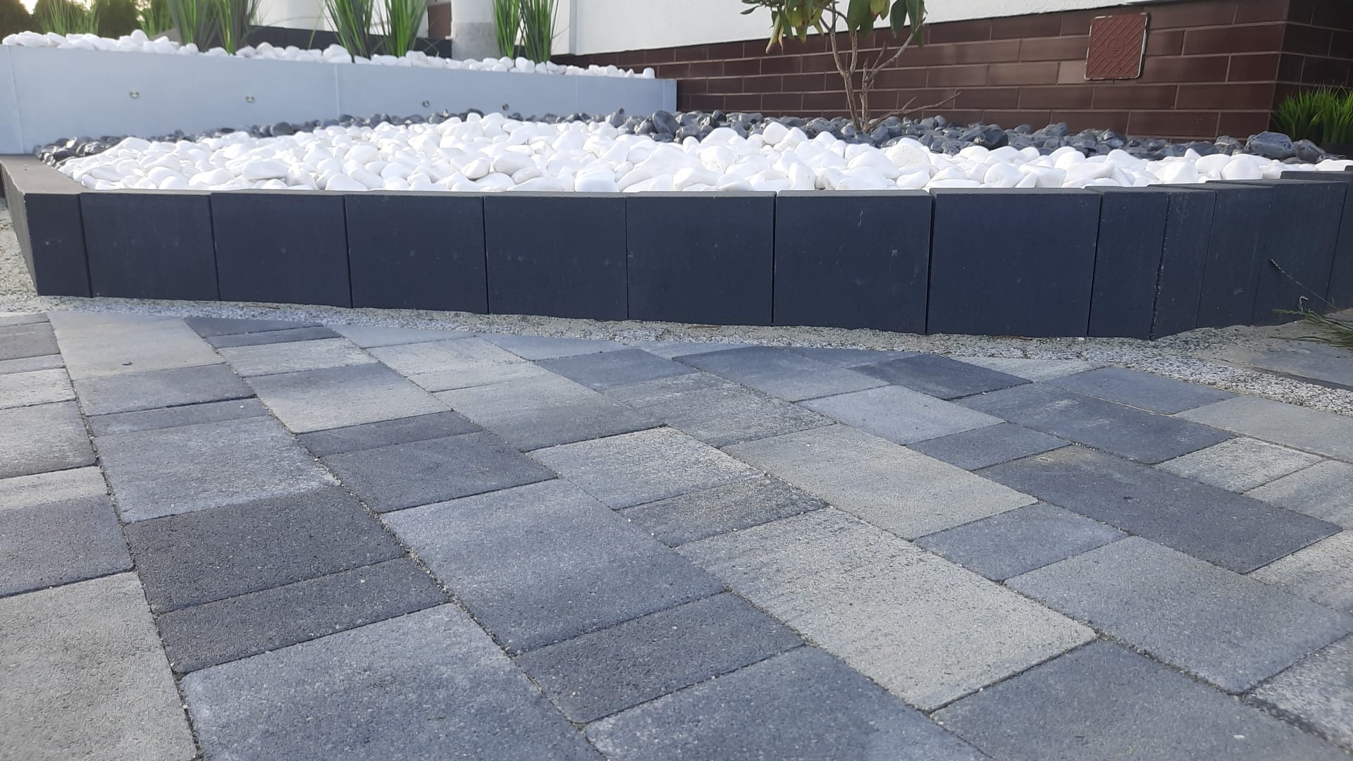 Ułożona szara kostka brukowa na podjeździe, obrzeża z ciemnych bloczków, rabata z białymi kamieniami i zielenią w tle.