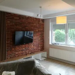 Salon w trakcie remontu: ściana z cegły klinkierowej, telewizor, zakryta folią kanapa, widoczny odkurzacz piorący.