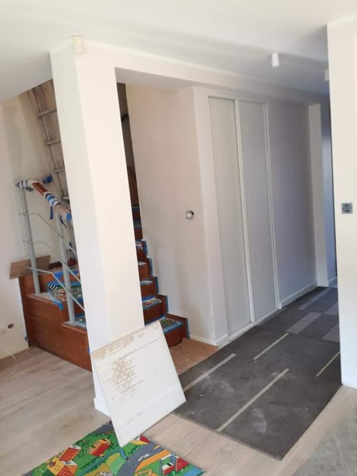 Wnętrze w trakcie remontu: drewniane schody zabezpieczone folią malarską, fragment korytarza z szarą wykładziną tymczasową, białe ściany po malowaniu, drabina oparta o ścianę, widoczne...