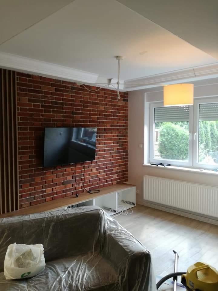 Salon w trakcie remontu: ściana z cegły klinkierowej, telewizor, zakryta folią kanapa, widoczny odkurzacz piorący.