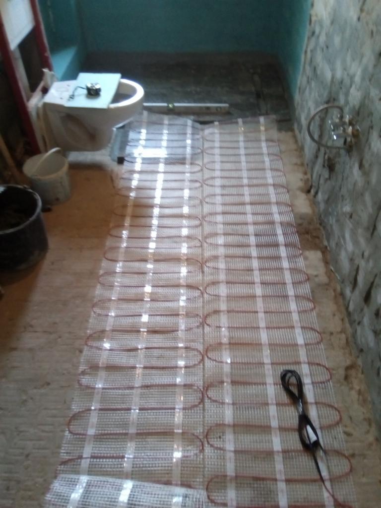 Mata grzewcza rozłożona na podłodze w trakcie montażu w łazience, widoczny fragment instalacji wodnej na ścianie i narzędzia.