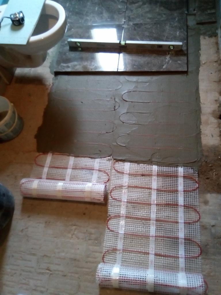 Montaż ogrzewania podłogowego: mata grzewcza rozłożona na podłożu z wylewką, widoczna poziomica na płytkach, przygotowania do instalacji w łazience.