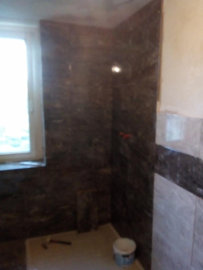 Fragment łazienki w trakcie prac wykończeniowych: ściany częściowo pokryte ciemnymi płytkami imitującymi kamień, widoczne narzędzia (szpachelka) oraz wiaderko z zaprawą na podłodze, okno w tle.