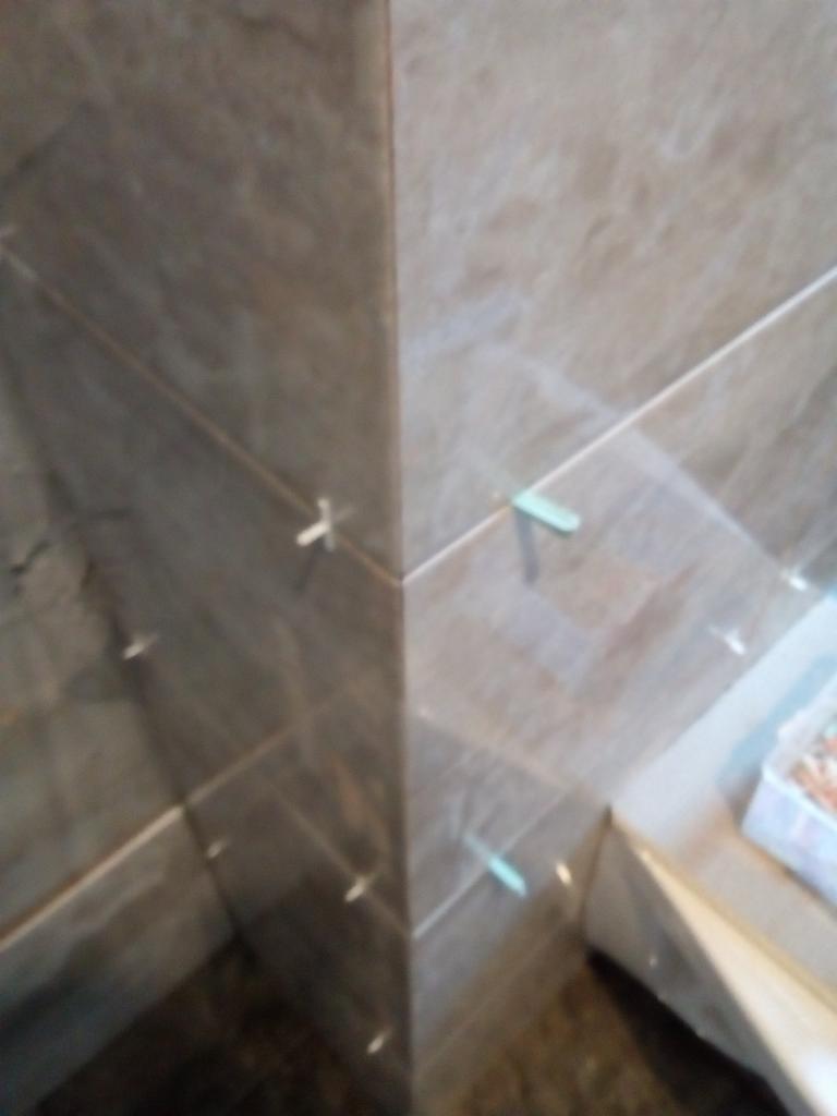 Fragment narożnika łazienki z ułożonymi płytkami imitującymi marmur, z widocznymi krzyżykami dystansowymi i pojemnikiem z zaprawą w tle.