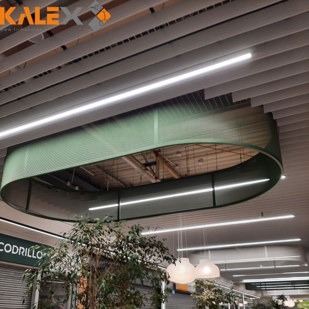 Zielona, perforowana konstrukcja podwieszona pod sufitem w przestrzeni handlowej, z widocznymi lampami liniowymi i roślinami w tle.