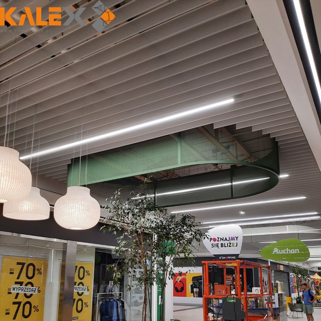 Równoległe, białe lamele sufitowe z wbudowanym oświetleniem liniowym w galerii handlowej, z zieloną, ażurową konstrukcją pod stropem i wiszącymi lampami kulistymi.