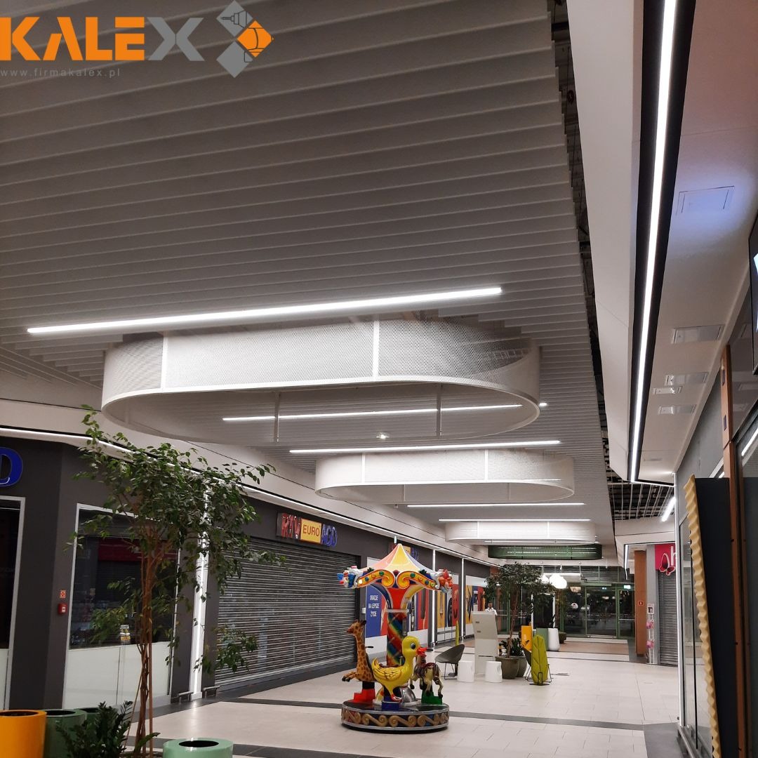 Sufit rastrowy z podwieszanymi, okrągłymi panelami akustycznymi i liniowym oświetleniem LED w przestrzeni handlowej z karuzelą dla dzieci.