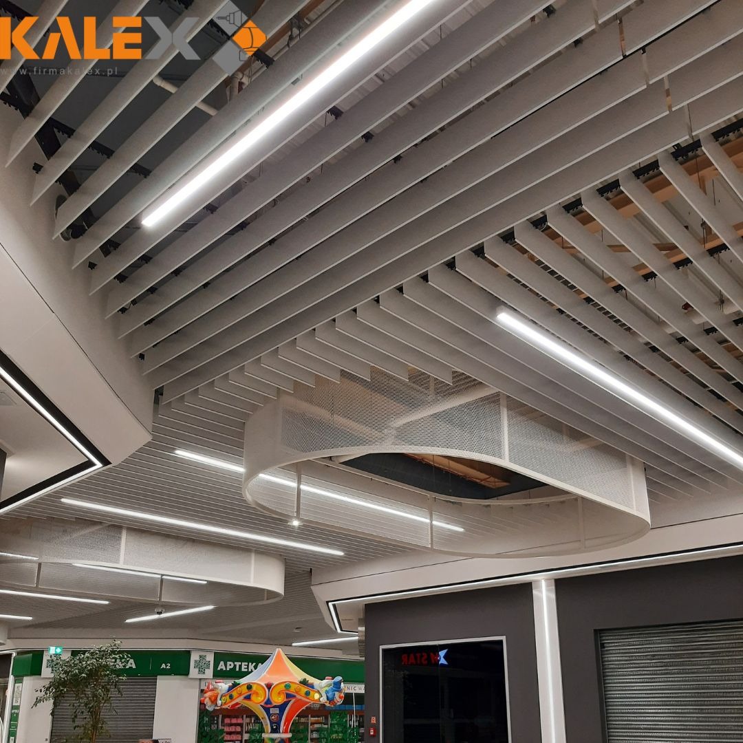 Biały sufit rastrowy z oświetleniem liniowym LED w centrum handlowym, widoczny fragment karuzeli i witryny sklepowe