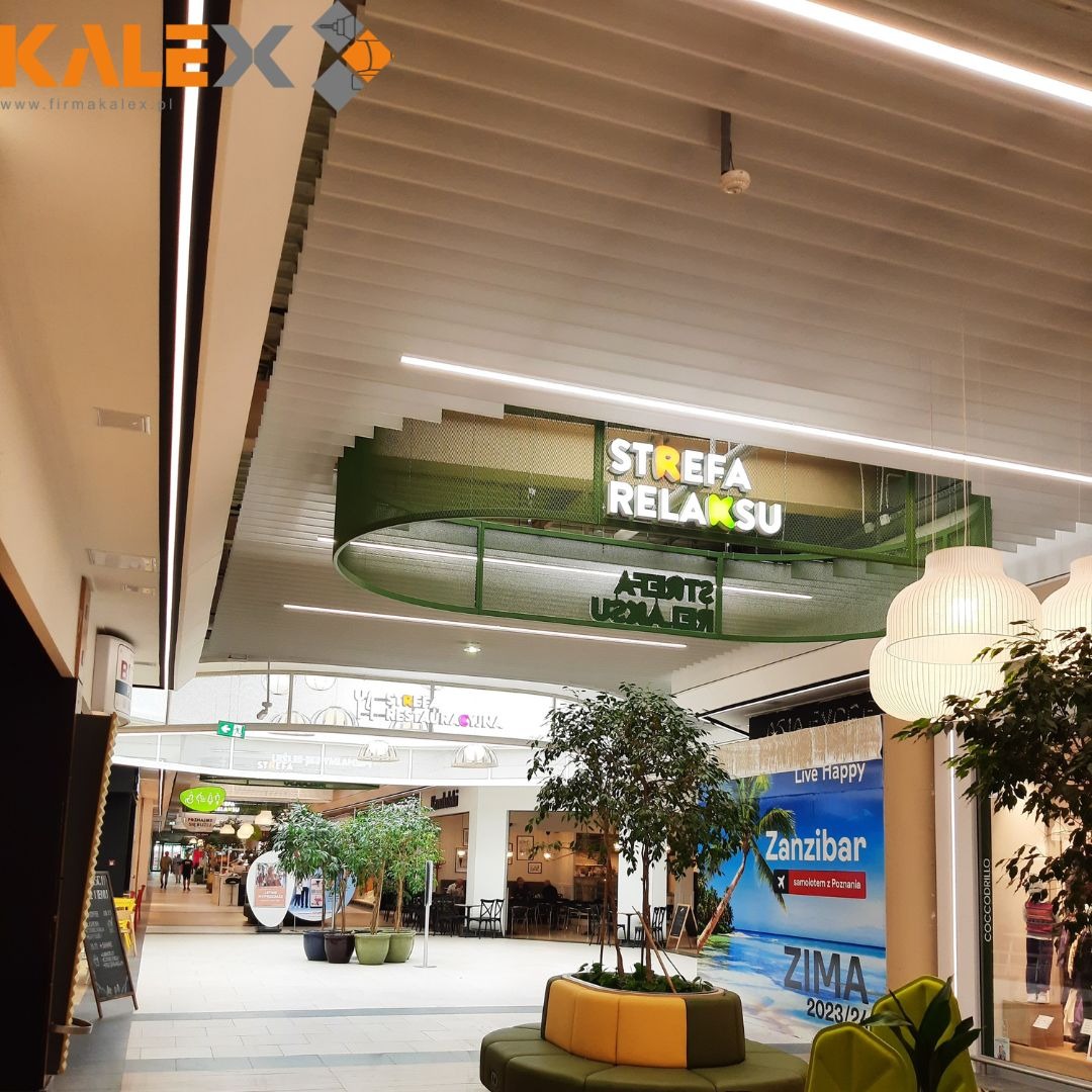 Wnętrze galerii handlowej z podwieszonym sufitem rastrowym, oświetleniem liniowym LED i dekoracyjną konstrukcją z napisem 'Strefa Relaksu' w kolorze zielonym, widoczne drzewka w donicach i plakat...