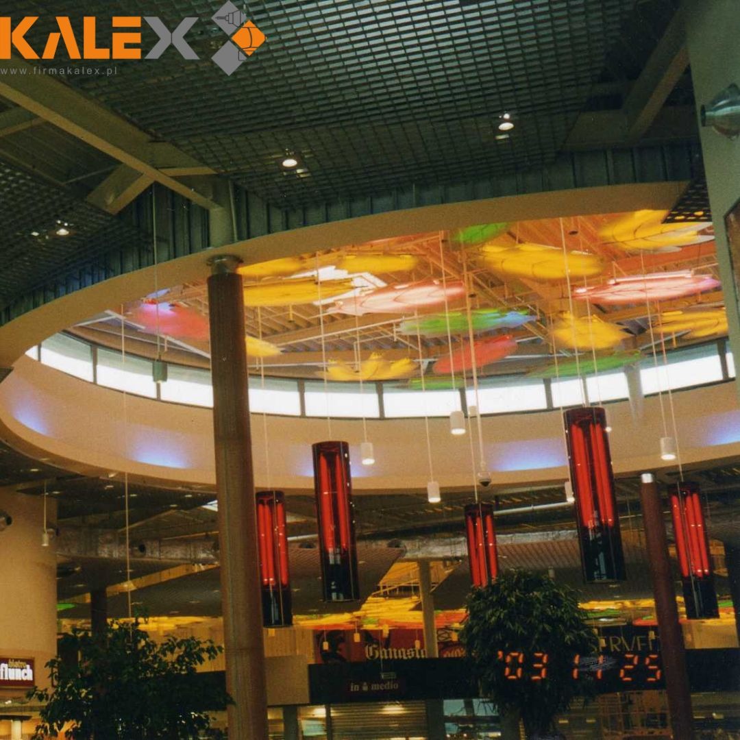 Sufit podwieszany z metalowej siatki w centrum handlowym, oświetlony kolorowymi panelami i pionowymi lampami, widok z dołu.