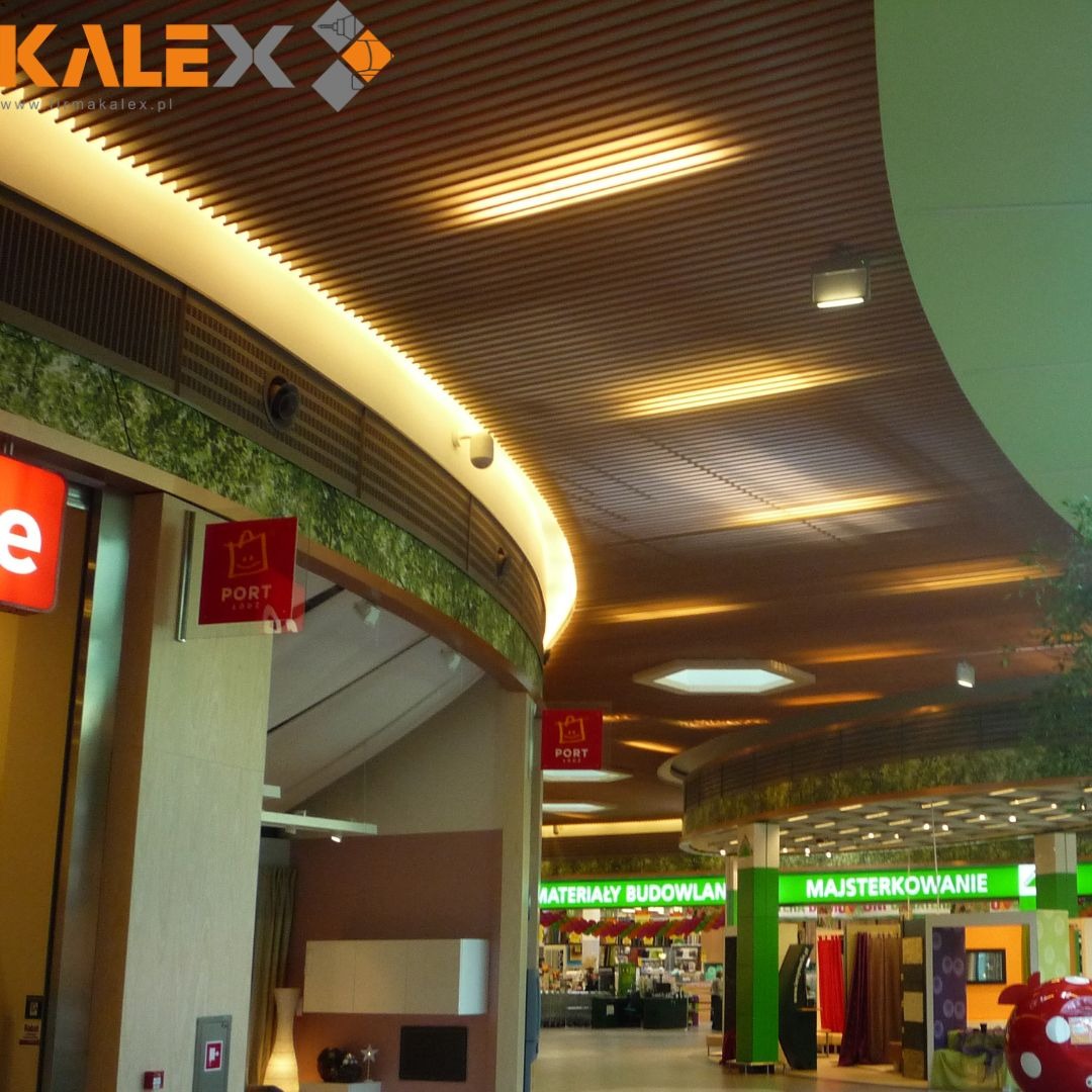 Wnętrze centrum handlowego z drewnianym, lamelowym sufitem podwieszanym z oświetleniem liniowym LED, widoczne sklepy z materiałami budowlanymi i majsterkowaniem.