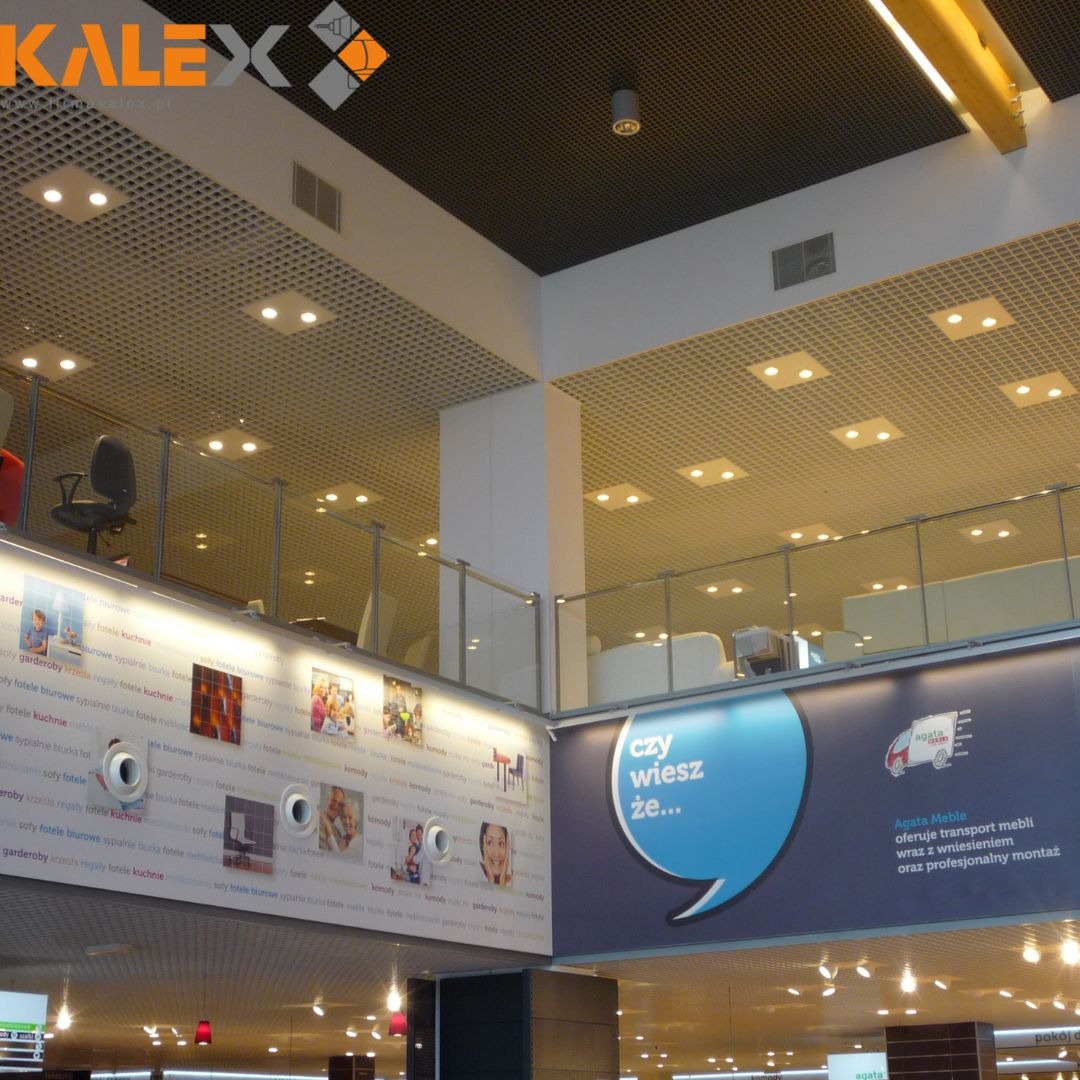 Sufit rastrowy z oświetleniem w przestrzeni biurowej lub handlowej, widok z dołu na konstrukcję sufitu podwieszanego z widocznymi elementami instalacji i reklamami na ścianach.