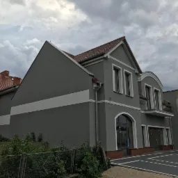 Szary, dwukondygnacyjny budynek z czerwonym dachem w Śmiglu, z białymi pasami i zdobieniami wok&oacute;ł okien, widziany z niskiej perspektywy pod pochmurnym niebem.