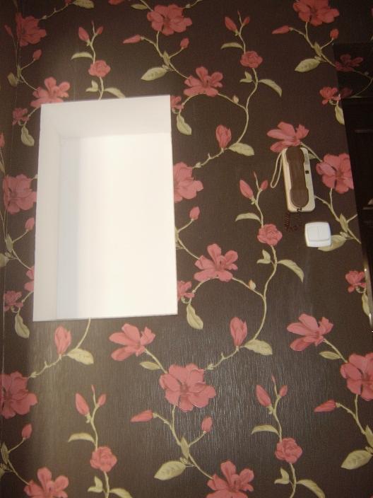 Ściana z ciemną tapetą w różowe kwiaty, wnęka z białym obramowaniem i zamontowany na ścianie telefon stacjonarny ze słuchawką i przyciskiem.