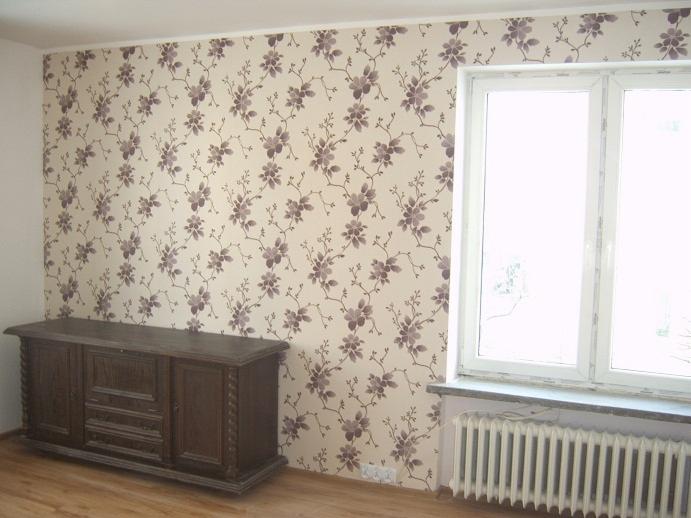Ściana z tapetą w drobne, fioletowe kwiaty, obok białe okno i grzejnik, na podłodze drewniana komoda.