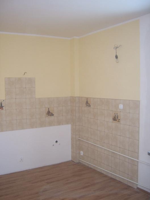 Wnętrze kuchni w trakcie remontu: częściowo położone beżowe płytki z motywem butelek, odsłonięta instalacja elektryczna i grzewcza, surowa żarówka zwisająca z sufitu.