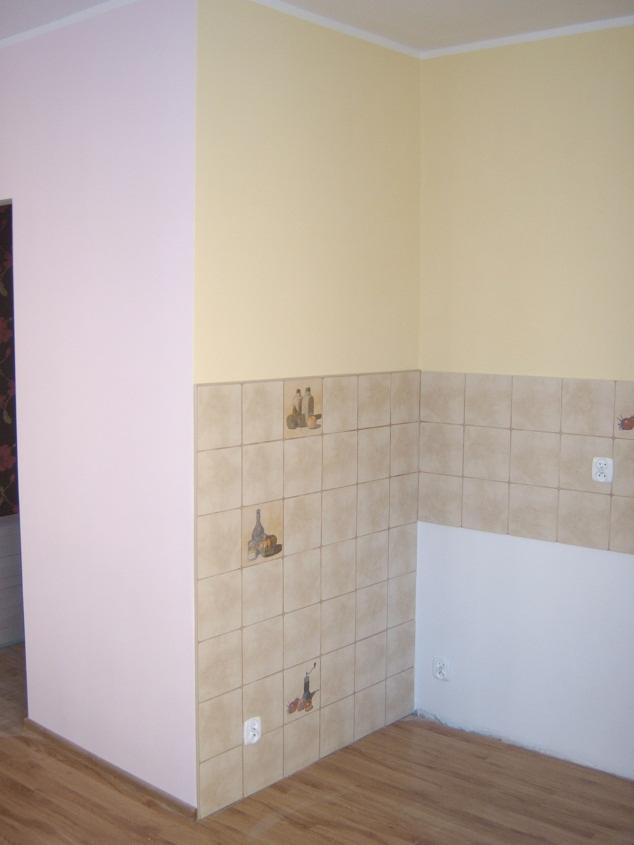 Świeżo pomalowane ściany w odcieniach różu i żółci w rogu pomieszczenia, z fragmentem ściany wyłożonej beżowymi płytkami z motywem butelek i owoców, oraz widocznymi gniazdkami elektrycznymi.