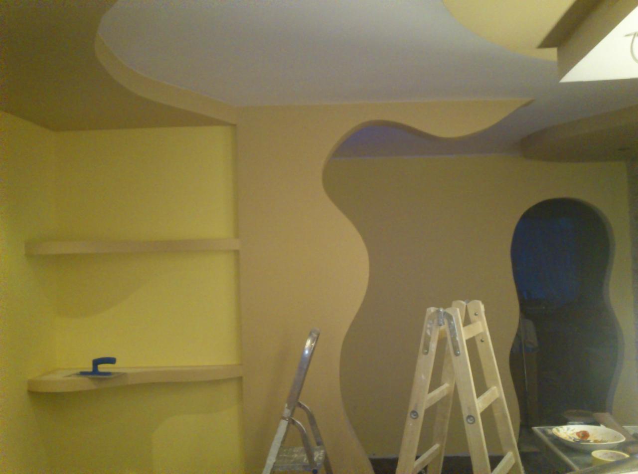 Żółte ściany z półkami i dekoracyjnym wycięciem w kształcie fali, widoczne dwie drabiny malarskie w trakcie prac wykończeniowych.