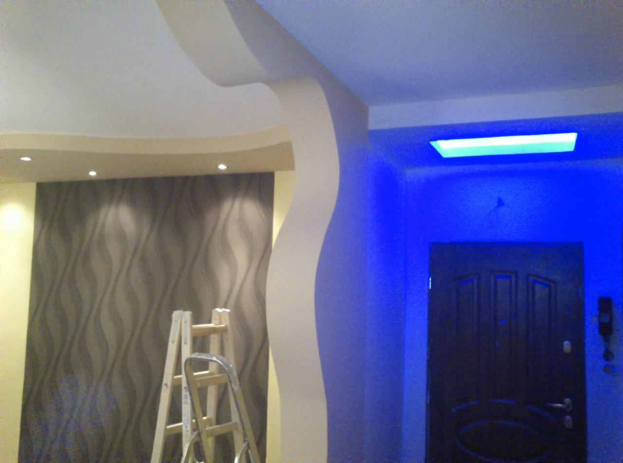 Wnętrze z dekoracyjną ścianą w odcieniach szarości, schodami malarskimi i podświetlanym sufitem oraz ścianą w kolorze intensywnego błękitu z ciemnymi drzwiami.