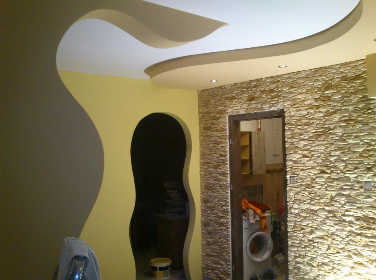 Aranżacja wnętrza z dekoracyjną ścianą z kamienia, nietypowym sufitem podwieszanym z oświetleniem punktowym i nieregularnym otworem drzwiowym w ścianie. W tle widoczna pralka i elementy wyposażenia...